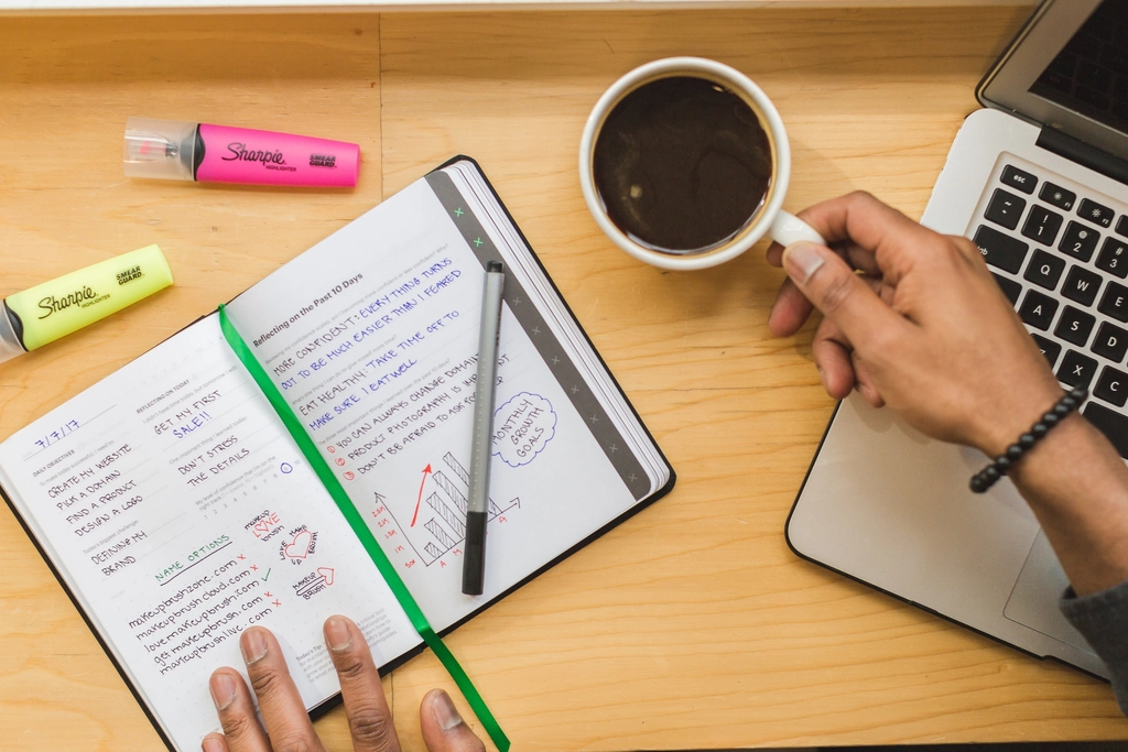 Mains posées sur un carnet ouvert à côté d'une tasse de café, ambiance de travail chaleureuse